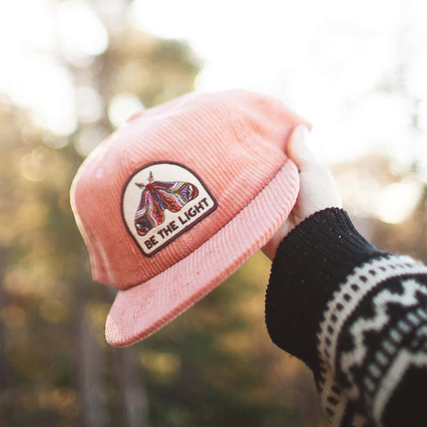 Person holding a coral Trek Light 6-panel snapback hat with a "Be The Light" moth design, flat brim, perfect for outdoor adventures.