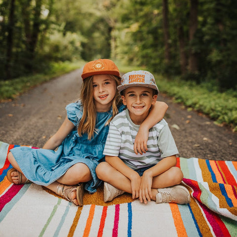 Two kids sitting on a colorful Trek Light blanket in the forest, wearing hats and smiling on a nature adventure.