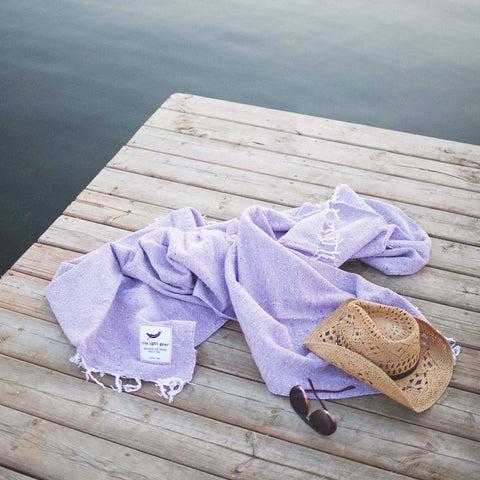 Lavender Sonoma Blanket, handwoven from recycled materials, on a dock with a straw hat and sunglasses, exuding cozy and versatile charm. By Trek Light.