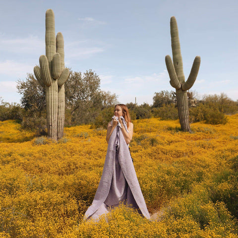 Woman wrapped in a lavender handwoven Sonoma Blanket, standing in a cactus-filled desert landscape, exuding beauty and coziness. By Trek Light.