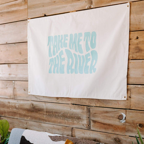 Trek Light's Take Me To The River Canvas Flag displayed indoors on a wooden wall, featuring bold text and brass grommets for hanging.