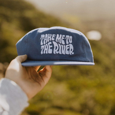 Hand holding a blue 'Take Me To The River' 6-panel snapback hat, perfect for river days, fishing, and adventures with Trek Light.