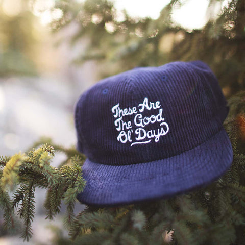 Trek Light navy hat with 'These Are The Good Ol' Days' embroidery, nestled on green pine branches, evoking nostalgia and warmth.