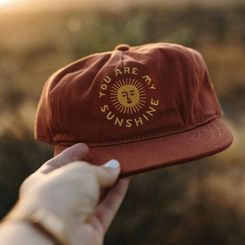 Hand holding a "You Are My Sunshine" 5-panel snapback hat by Trek Light, featuring a flat brim and embroidered sun design.