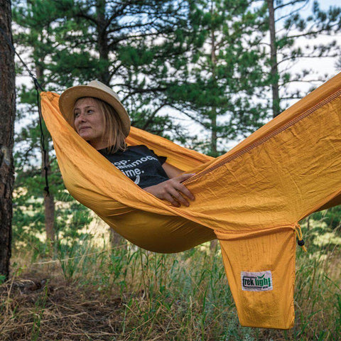 Person relaxing in a yellow Trek Light banana hammock in a forested area, enjoying nature and supporting cancer awareness.