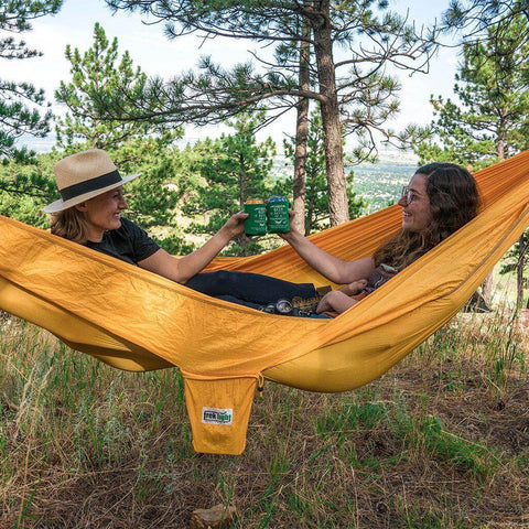 Two people relaxing in Trek Light Banana Hammock, raising mugs amidst forested scenery.