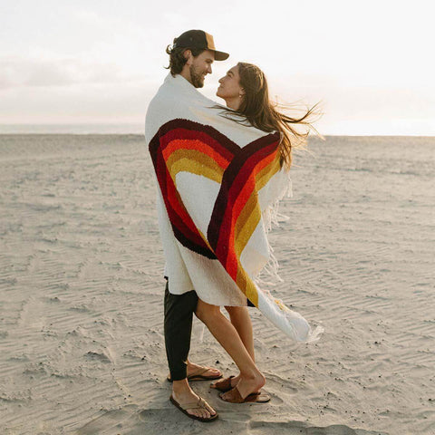 Couple wrapped in Trek Light's Bowie Blanket with vintage rainbow design, enjoying a beach moment.