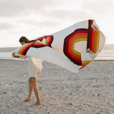 Person holding vintage rainbow Bowie Blanket on a beach, showcasing eco-friendly Trek Light design, handwoven with recycled materials.