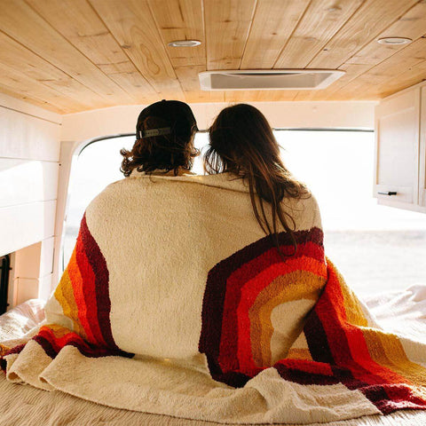 Couple wrapped in a Trek Light Bowie Blanket with rainbow design, enjoying a cozy moment in a wooden van interior.