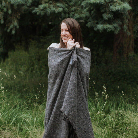 Smiling person wrapped in a beautiful, handwoven grey Trek Light blanket made from recycled clothing materials, showcasing its cozy versatility.