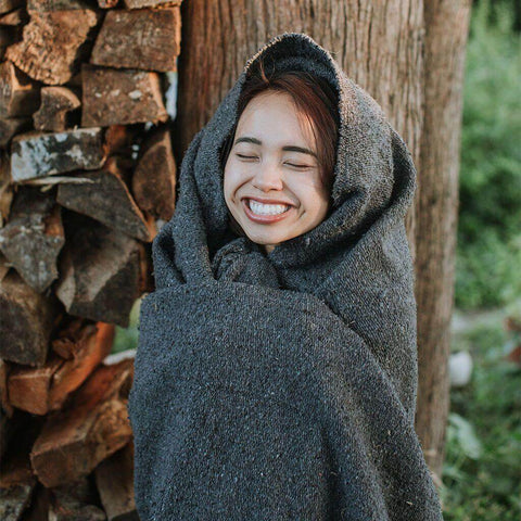 Woman joyfully wrapped in a cozy Trek Light Greyson Blanket, handwoven from recycled clothing materials, showcasing its versatile beauty.