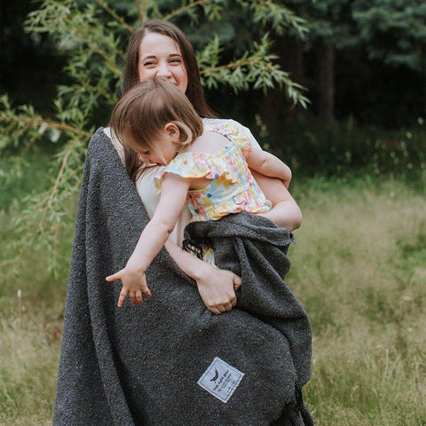 Woman holding a child wrapped in a cozy Trek Light Greyson Blanket, handwoven from recycled materials, embodying comfort and playfulness.
