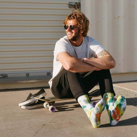 Man wearing Happiest Camper Tie Dye Socks by Trek Light, sitting with a skateboard, enjoying the sun and ready for adventure.