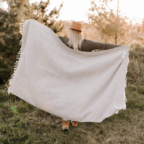 Woman holding a sand-colored Huntington Blanket in nature, perfect for beach days and cozy nights. Trek Light adventure vibes.