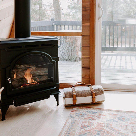 Cozy Trek Light blanket rolled with artisan leather carrier beside a warm fireplace, ready for stylish adventures.