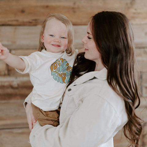 Child wearing 'Love Your Mother' Trek Light shirt, smiling in mother's arms. Eco-friendly message, soft cotton tee for happy families.