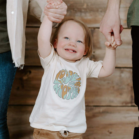 Happy child wearing 'Love Your Mother' Trek Light kids shirt, held by parents, showcasing eco-friendly design and family love.