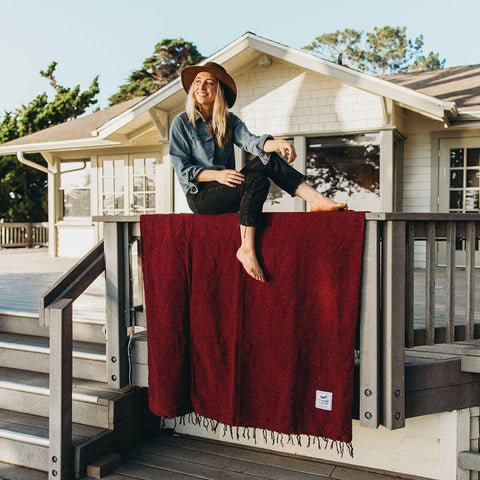 Woman in a hat sitting on a porch railing above a red Trek Light Napa Blanket, made from recycled materials. Eco-friendly cozy vibes.