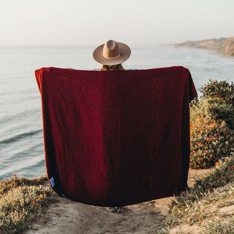 Person wrapped in red Napa Blanket by the ocean, cozy and eco-friendly, perfect for naps or enjoying nature. Trek Light.
