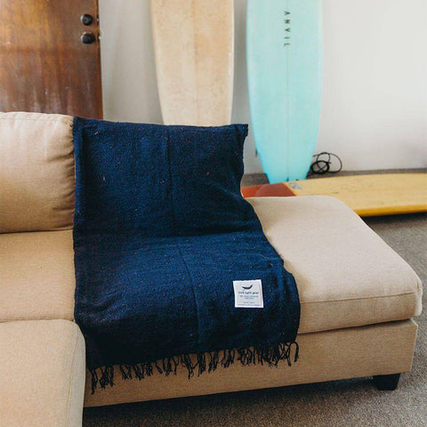 Navy Newport Blanket from Trek Light draped over a beige couch, with surfboards in the background, embodies coastal comfort.