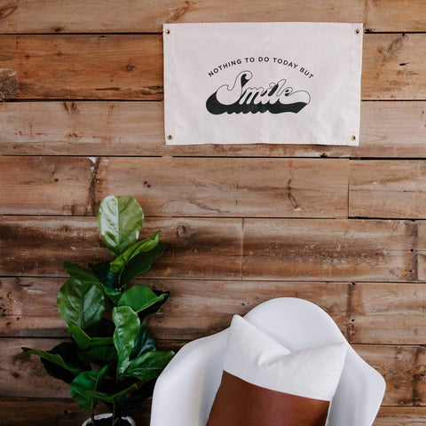 Trek Light 'Nothing To Do Today But Smile' canvas flag on rustic wood wall with chair and plant; 16x26 inch flag in natural cotton.