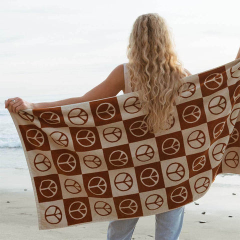 Person holding a Trek Light peace-sign patterned blanket on a beach, ocean in the background.