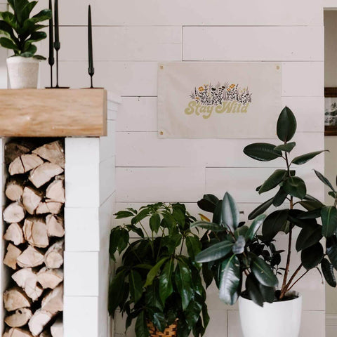 Stay Wild Canvas Flag by Trek Light hanging on a white wall with indoor plants, featuring hand-screened art and brass grommets.