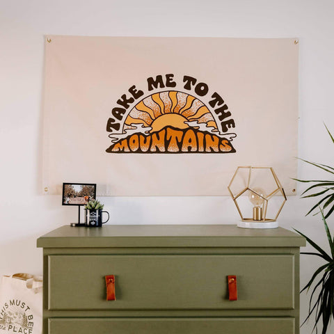 Trek Light's "Take Me To The Mountains" canvas flag with sunrise design, hanging above a green dresser. Perfect for mountain lovers.