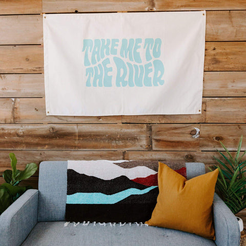 Trek Light Take Me To The River Canvas Flag hanging above a cozy couch with cushions, showcasing artistic home decor.