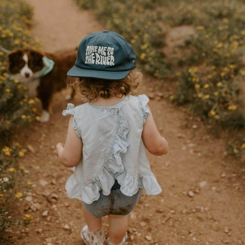 Adorable child wearing 'Take Me To The River' hat by Trek Light, exploring a scenic path with a cute puppy nearby.