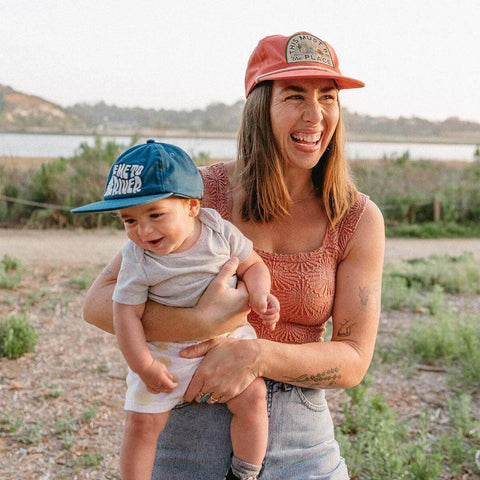 Smiling woman and baby outdoors, both wearing snapback hats. Baby in "Take Me To The River" kids hat by Trek Light, ready for adventure.