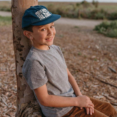 Young boy wearing Trek Light 'Take Me To The River' blue kids hat, smiling under a tree, ready for an outdoor adventure.