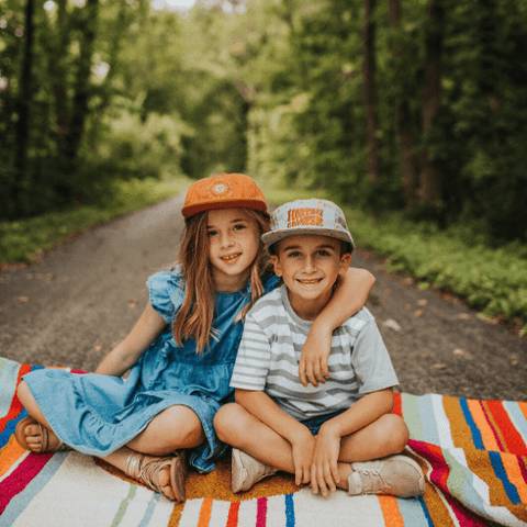 Two smiling kids wearing Trek Light hats sitting on a colorful blanket outdoors on a forest path.