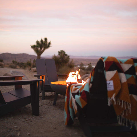 Beautiful handwoven Cascade Blanket made from recycled materials, draped over a cozy chair by a fire in a tranquil desert setting. By Trek Light.