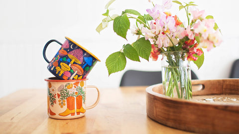 Decorative Trek Light enamel mugs with colorful designs stacked beside a vase of fresh flowers on a wooden table.