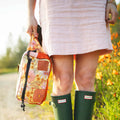 Woman outdoors wearing green boots, holding a floral Trek Light Everyday Fanny Pack. Perfect for style and adventure.
