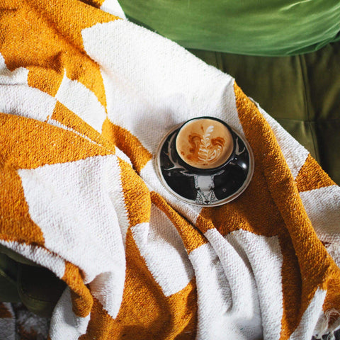 Cozy handwoven Goldie blanket in geometric design, made from recycled clothing materials, with a cup of coffee on top. By Trek Light.