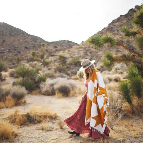 Woman walking in desert with Goldie Blanket, a beautiful, handwoven, and versatile throw made from recycled clothing materials. By Trek Light.