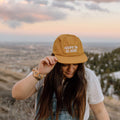 Happy To Be Here hat by Trek Light, worn by a smiling person with scenic landscape background, 6-panel unisex snapback.