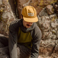 Man wearing 'Happy To Be Here' yellow snapback hat sitting against rocky backdrop, featuring stylish vent holes. Trek Light brand.