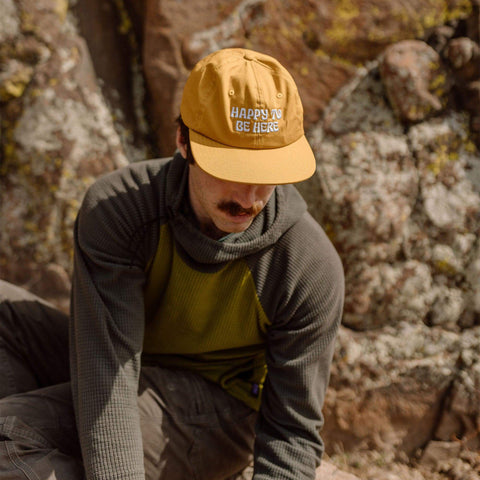 Man wearing 'Happy To Be Here' yellow snapback hat sitting against rocky backdrop, featuring stylish vent holes. Trek Light brand.