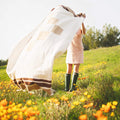 Person joyfully holding a cozy, handwoven North Star Blanket made from recycled clothing materials in a vibrant flower field. By Trek Light.