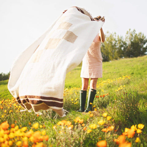 Person joyfully holding a cozy, handwoven North Star Blanket made from recycled clothing materials in a vibrant flower field. By Trek Light.