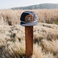 Trek Light Mountains Hat on a wooden post in a field, ready for adventure. This Must Be The Place snapback with flat brim.