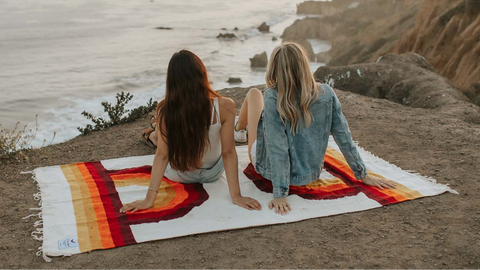 Two friends enjoying an outdoor adventure on a colorful Trek Light blanket by the ocean, embracing a scenic sunset view.