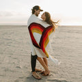 Couple wrapped in Trek Light's Bowie Blanket with vintage rainbow design, enjoying a beach moment.