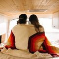 Couple wrapped in a Trek Light Bowie Blanket with rainbow design, enjoying a cozy moment in a wooden van interior.