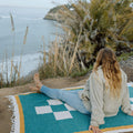 Woman relaxing on a Trek Light Dolly Blanket by a coastal view, embracing nature and comfort with sustainable style.