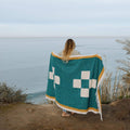 Woman standing by the ocean wrapped in a Trek Light Dolly Blanket, made from recycled materials, showcasing a nature-friendly design.
