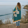 Person wrapped in a Trek Light Dolly Blanket overlooking the ocean, showcasing eco-friendly, handwoven design.